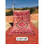 Un Tapis berbère tissé main en laine rose et noir tissé à la main | 343x186 cm posé sur un tapis rouge et noir.